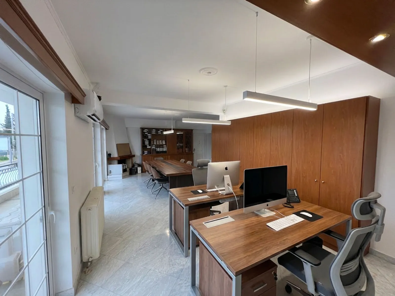 Bright, open office with a long wooden conference table, ergonomic chairs, and tall wooden storage cabinets along the wall.