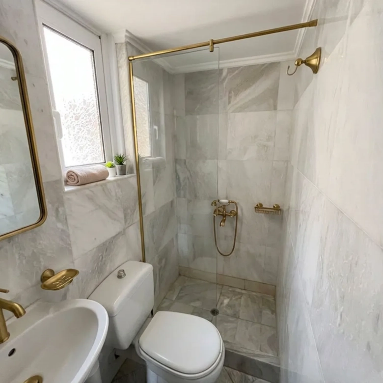 Compact marble-tiled bathroom with gold fixtures, glass shower enclosure, sink, and toilet; towel and small plants on the frosted window ledge.