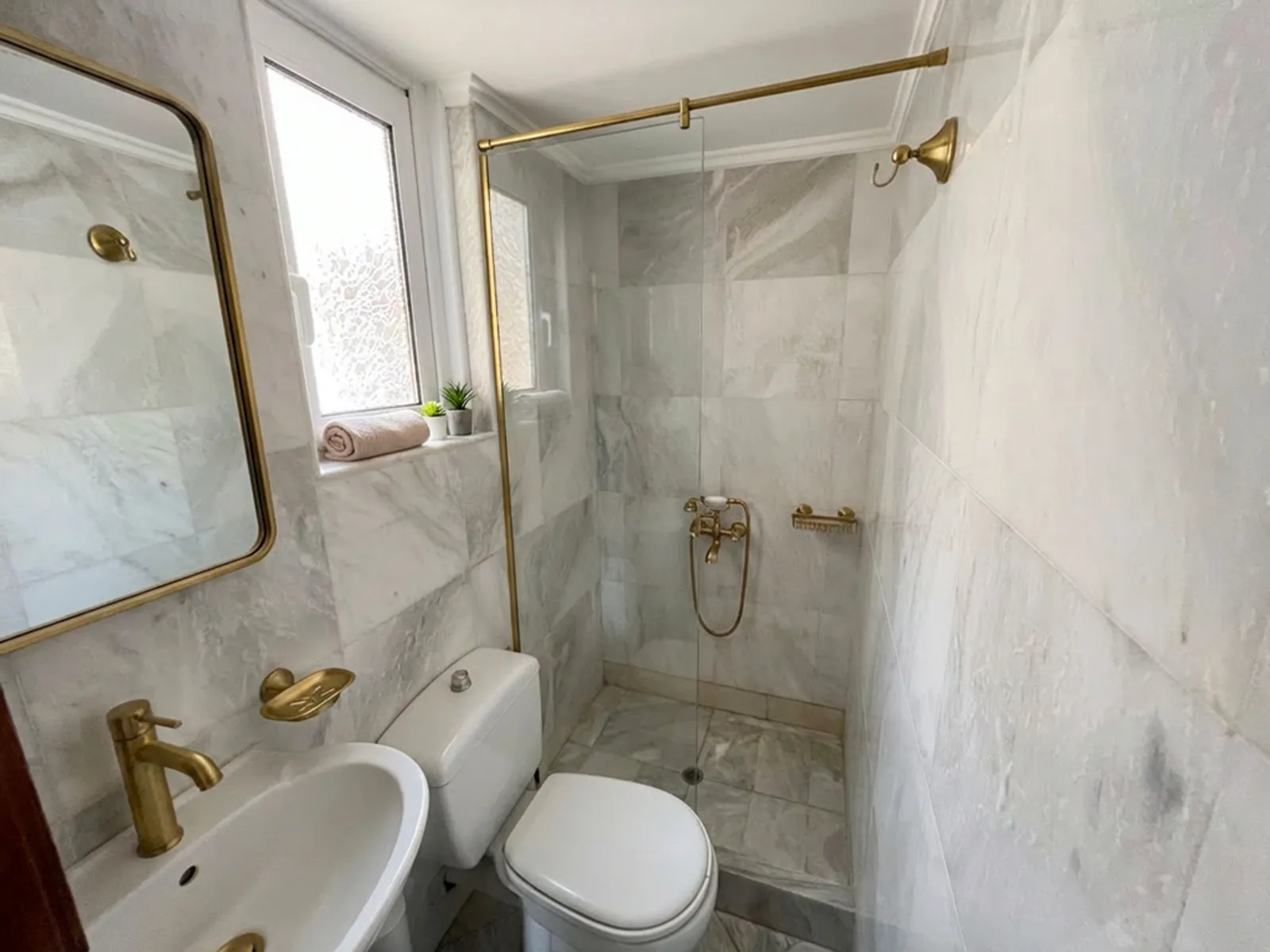 Compact marble-tiled bathroom with gold fixtures, glass shower enclosure, sink, and toilet; towel and small plants on the frosted window ledge.