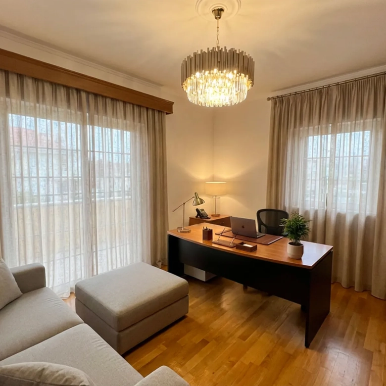 Cozy home office in a living room: wooden desk with laptop, chair, desk lamp, and a chandelier overhead; beige sheer curtains and a gray sofa nearby.