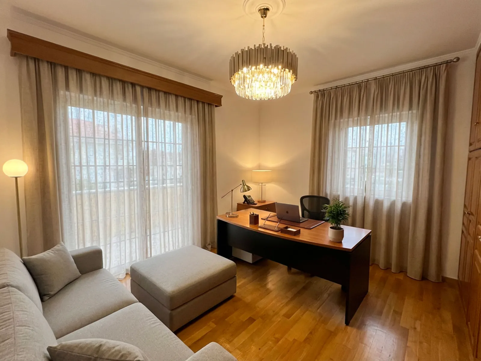 Cozy home office in a living room: wooden desk with laptop, chair, desk lamp, and a chandelier overhead; beige sheer curtains and a gray sofa nearby.
