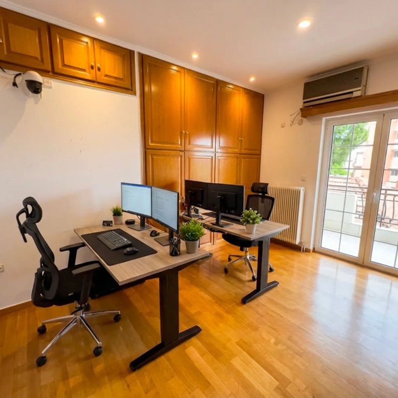 Home office with two desks, dual monitors, and plants near a glass door to a balcony.