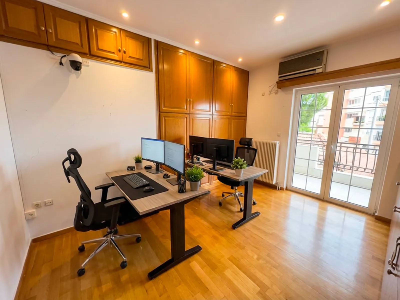 Home office with two desks, dual monitors, and plants near a glass door to a balcony.