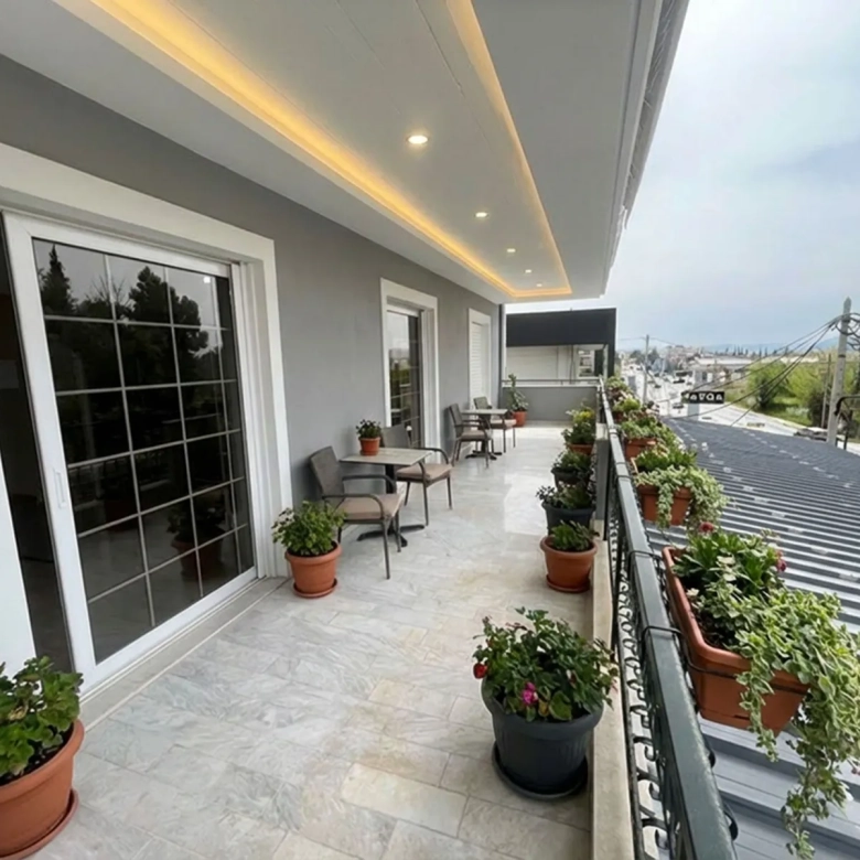 Balcony with potted plants, seating, and sliding glass doors along a gray building.