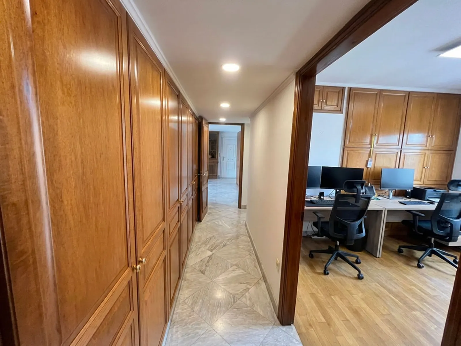 Long hallway with tall wooden cabinets lining the left wall and an open office area with desks and computers on the right.